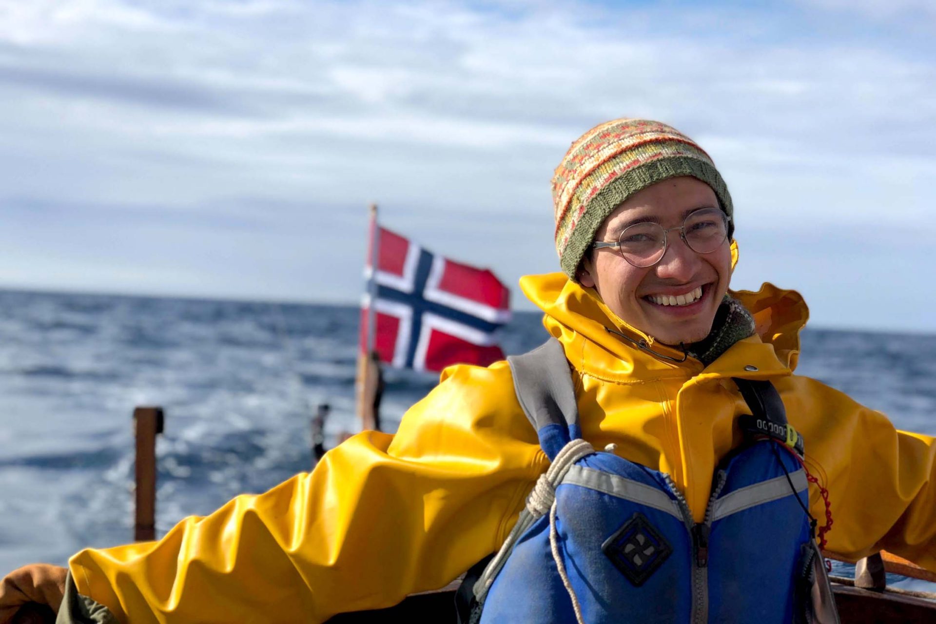 Seiling-Fosen-Folkehogskole-Styrer-sol-seilbåt En glad elev med regntoy og flytevest styrer seilbaten i frisk vind. Det norske flagget i bakgrunnen.